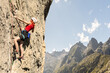 © VICTOR - A woman is climbing a rock wall with a red shirt and blue shorts. The mountain in the background is covered in trees