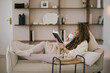 © polinaloves - Young woman reading book and drinking tea, sitting on sofa in a cozy living room.