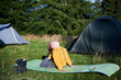Young boy engaged in yoga pose on green mat in grassy field in the camping. Two tourist tents are set up among a copse of green trees on background.