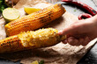 Caucasian hand holds half eaten grilled corn on cob with lime and other spices on wooden table