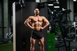 Dark-haired bodybuilder performing front lat spread pose in gym. Front view of strong Caucasian male athlete in shorts, lifting chest up, spreading lats and showing to camera. Concept of bodybuilding.