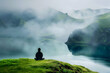 Someone sitting in a lotus position on a grassy hill, facing a calm lake with morning fog hovering over the water