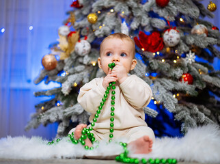 Wall Mural - A baby is sitting on the floor in front of a Christmas tree. The baby is holding a green string in its mouth