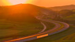 AERIAL, LENS FLARE: Sun slowly setting over a winding road and scenic landscape. Evening sunbeams cast warm orange hues on the countryside intersected by the highway that meanders past rolling hills.