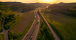 AERIAL, LENS FLARE: Smooth running traffic on a motorway winding through hills and lush green countryside. Cars and trucks travel along the highway as morning light illuminates picturesque landscape.