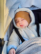 A baby is sleeping in a stroller with a brown hat on. The baby is wearing a white jacket and a blue shirt