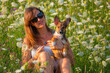 PORTRAIT, DOF: Woman lovingly holds her small puppy amidst a field of blooming wildflowers. Smiling lady and her adopted doggo have a photo session on a meadow in warm embrace of golden sunlight.