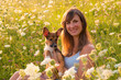 PORTRAIT, DOF: Young lady poses with an adorable puppy amidst lush wildflowers. Woman holds her adopted doggo as they sit surrounded by the natural beauty of a blossoming meadow in golden sunlight.