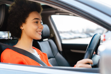 Smiling attractive young African American woman with curly hair driving car, buckled safety belt