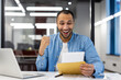 © Tetiana - Smiling young hispanic man sitting in modern office and reading letter received in mail, happy with news and message