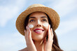 Beautiful Young woman with sun cream on face. Girl holding sunscreen bottle on the beach. Female in hat applying moisturizing lotion on skin.Skin care. Sun protection. Suntan