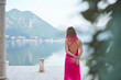 A woman with pink hair in a flowing dress stands facing a breathtaking lakeside view, with a Shiba Inu looking towards her