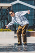 Young Man In Stylish White Marble Pattern Sweatshirt And Brown Pants Performing A Dynamic Dance Move In An Urban Setting With Modern Glass Buildings In The Background. Vertical Screen.