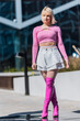 Vertical Screen. Striking Image Of A Young Blonde Woman In A Pink Crop Top And White Skirt, Paired With Pink Boots, Walking Confidently In An Urban Setting With Modern Architecture In The Background.