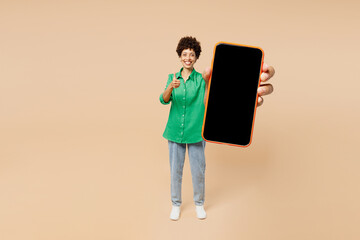 Wall Mural - Full body young woman of African American ethnicity wear green shirt casual clothes hold in hand use blank screen mobile cell phone show thumb up isolated on plain beige background. Lifestyle concept.