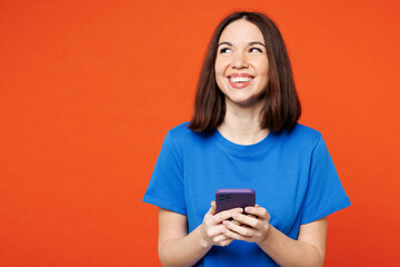 Wall Mural - Young fun surprised happy woman she wear blue t-shirt casual clothes hold in hand use mobile cell phone look aside on area isolated on plain red orange background studio portrait. Lifestyle concept.