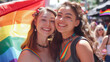 © Anthichada - Happy smiling young woman lesbian couple with lgtbq flags celebrating gay pride month together. Lesbian couple raising a rainbow flag at a gay pride parade celebrating pride month. LGBTQ+, Lesbian