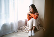 Pensive young Asian woman in warm knitted clothes sitting on windowsill in room. Negative human facial expression.