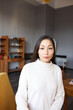 Vertical portrait of pretty female psychotherapist sitting in chair in her cozy cabinet before visit of her client. Adorable asian woman waiting for her therapist sitting in lobby looking at camera