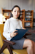 Worried anxious elegant asian female in casual skirt and sweater sitting in armchair in stylish office with clipboard and pen, filling in blank before job interview on desired vacant position