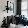 Close-up of dark gray headboard with many pillows, poster in a black frame and sconce on white wall next to the window with blinds and gray curtains, green glass bottle, and decor on a bedside table.