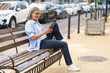 A woman is sitting on a bench and looking at her tablet. She is smiling and she is enjoying her time. The scene takes place on a street with several cars parked nearby