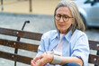 A woman sits on a bench with her hands on her knees. She is wearing glasses and a blue shirt