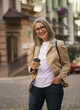 A woman in a tan jacket and glasses is smiling while holding a coffee cup. She is wearing a white shirt and blue jeans
