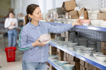 Wall Mural - Female shopper choosing with interest ceramic bowls for udon and ramen among variety of dishware arranged on shelves at Asian store..