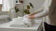 A person neatly folding clean laundry on a simple white table in a white room, conveying a sense of domestic routine. 