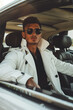 Confident stylish male model in white trench coat and sunglasses sitting in a luxury car