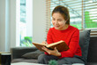 © Prathankarnpap - Peaceful young woman in red sweater reading book on comfortable couch at home