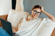 © SHOTPRIME STUDIO - Woman using laptop on a cozy couch at home freelancer in a modern home office She's surrounded by technology, enjoying the online lifestyle With a smile and glasses on, she works comfortably and