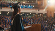 © Dina Photo Stories - Young happy woman in a gown and a mortarboard stands at a podium and gives a graduation speech. Valedictorian young female student wearing graduation hat giving graduation speech to the audience