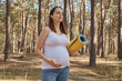 Smiling pregnant woman wearing casual clothing standing in forest enjoying nature after workout holding yoga mat looking away with pensive facial expression