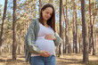 Beautiful pregnant woman wearing green jumper stroking her belly with hands smiling with love breathing fresh air in woodland on last months of pregnancy