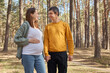 Cheerful young family pregnant wife and her husband holding hands while walking in forest enjoying communicating spending weekend together during pregnancy
