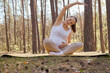 Smiling adult Caucasian pregnant woman doing yoga on mat stretching her hands and sides while sitting in lotus pose enjoying sport workout in woodland