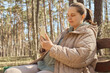 Brown haired woman with ponytail texting on the smart phone while sitting in park wearing jacket and sitting in bench in park browsing internet in open air