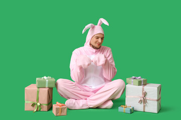 Canvas Print - Young man in bunny costume with gift boxes sitting on green background. Easter celebration