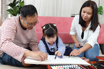 Happy Asian family. Chubby little girl daughter drawing together with father and mother in living room. Parents spend time with toddler kid at home. Child, mom and dad playing together in house.