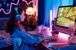 © AnnaStills - Modern teen girl wearing headphones sitting at desk in her bedroom decorated with neon lights playing video game on desktop computer