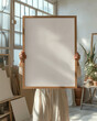 © Amy - A woman female artist holding a blank wood oak thin framed 20x28, 50x70 art print mockup, in a plant filled artist studio, with dappled sunlight from window