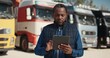 Attractive African American male driver using his work tablet device while standing before trucks. Planning road route for work trip. International transfer. Transportation. Concept of shipment.