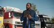 Attractive African American male driver using his work tablet device while standing before trucks. Planning road route for work trip. International transfer. Transportation. Concept of shipment.