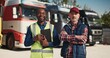 Mixed-race men working together at delivery business company. Attractive coworkers wearing cap standing before lorries with goods and positively smiling on camera. Transport concept.