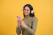 © Jose Calsina - Smiling happy teenage woman using her smartphone and chatting online or browsing on internet at yellow wall background. Young caucasian female watching funny videos with mobile phone application