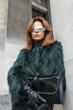 Cool fashion pretty lady with red hair with stylish sunglasses in fur jacket with bag stands near a vintage building