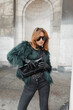 Fashion beautiful urban vogue model woman with sunglasses in a stylish shaggy jacket with a handbag posing in the city near the building