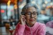 Elderly gray haired Latin American female in glasses and pink sweater sitting at table adjusting earphones while listening to audio on mobile phone